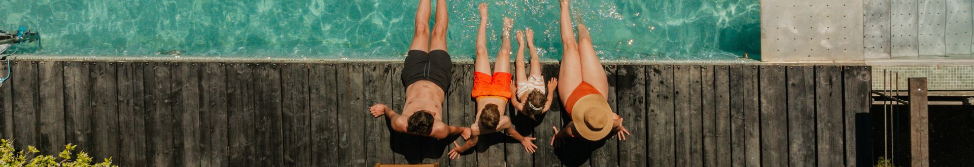 Draufsicht auf eine Familie am Poolrand: Eltern und zwei Kinder sitzen auf einem Holzsteg und lassen die Füße ins türkise Wasser baumeln