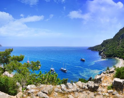 Blick auf die Bucht von Olympos bei Çıralı in der Türkei mit kristallklarem Wasser, mehreren Segelbooten und dicht bewaldeten Berghängen unter leicht bewölktem Himmel