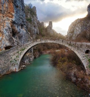 Steinerne Bogenbrücke über grünes Flusswasser in der Vikos-Schlucht, umgeben von steilen Felswänden und bewaldeten Hängen.