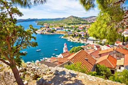 Blick über die Küstenstadt Šibenik in Kroatien mit roten Dächern, Kirchturm, Booten im Hafen und der Adriaküste zwischen grünen Hügeln