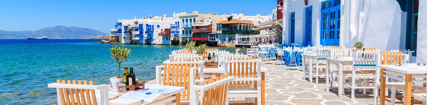 Freilufttaverne mit weißen Holztischen und Stühlen auf gepflastertem Weg am Meer vor weiß-blauen Gebäuden unter blauem Himmel