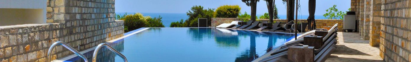Infinity-Pool zwischen Natursteinmauern mit Liegestühlen und Sonnenschirmen am Rand und Meerblick im Hintergrund