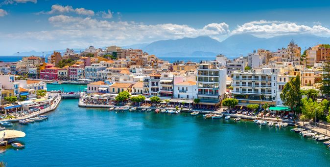 Blick auf den Hafen von Agios Nikolaos auf Kreta mit bunten Gebäuden, Booten und Bergen im Hintergrund.