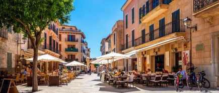 Straßencafé in der Altstadt von Alcúdia auf Mallorca mit historischen Häusern, Sonnenschirmen und Gästen im Freien