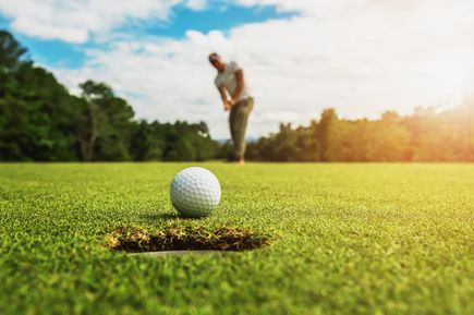 Nahaufnahme eines Golfballs kurz vor dem Loch, im Hintergrund unscharf ein Golfer beim Putten auf einem grünen Platz