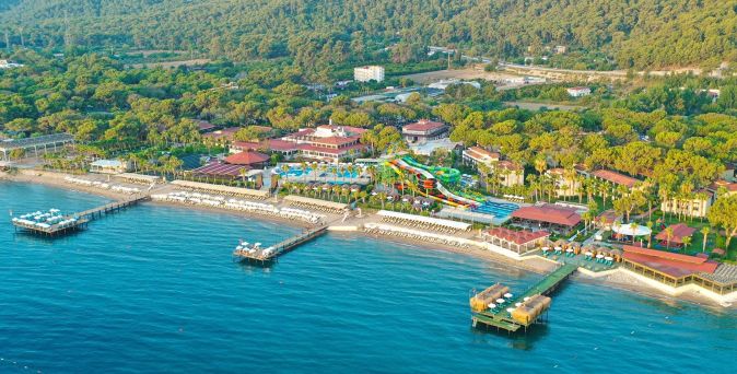 Luftaufnahme eines Crystal Hotels mit mehreren Gebäuden, Wasserrutschen, Liegestühlen und Stegen am Meer, umgeben von Wald und Bergen