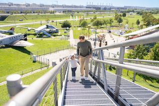 Erwachsener und Kind gehen eine Treppe im Besucherpark des Flughafens München hinauf, im Hintergrund historische Flugzeuge, Grünflächen und Spielbereiche für Besucher