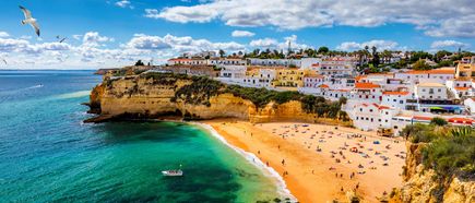Blick auf den Strand von Carvoeiro an der Algarve mit goldenen Felsen, türkisfarbenem Meer, Badegästen, weiß getünchten Häusern auf den Klippen und einer Möwe über der Küste