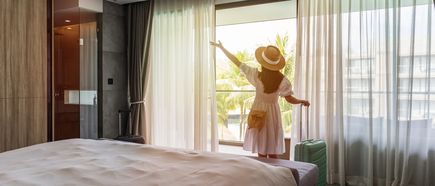 Frau mit Hut und weißem Kleid steht in Hotelzimmer vor großem Fenster und blickt nach draußen