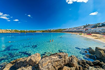 Blick auf die Bucht von Arenal d'en Castell auf Menorca mit klarem türkisblauem Wasser, Badestrand und weißen Ferienanlagen am Hang