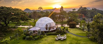 oderner Glamping‑Dome in grüner Landschaft bei Sonnenuntergang, umgeben von Bäumen, Wiesen und Hügeln, Sitzgruppe im Garten, Natur‑ und Erholungsatmosphäre