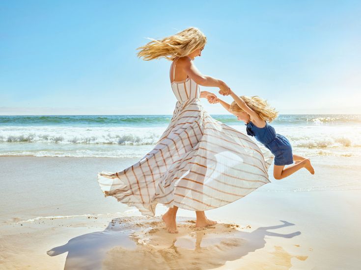 Frau schwingt ein Kind am Strand am Meer, beide barfuß im Sand, Wellen im Hintergrund, sonnige Strandlandschaft unter blauem Himmel