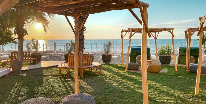 Strandbereich mit Holzpergolen, Sitzbänken und Sonnenschirmen am Meer bei Sonnenaufgang.
