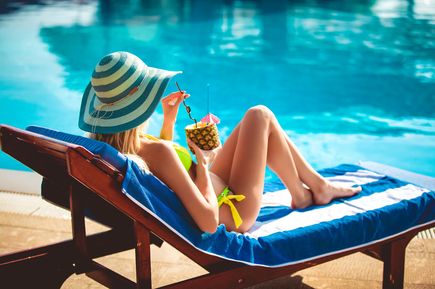 Frau mit gestreiftem Sonnenhut und gelbem Bikini entspannt auf Sonnenliege am Pool und hält Ananas-Getränk mit Strohhalm und kleinem Schirmchen