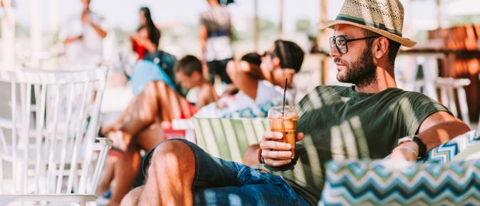 Mann mit Strohhut und grünem T-Shirt sitzt entspannt auf einer Terrasse und hält ein Getränk mit Strohhalm in der Hand.