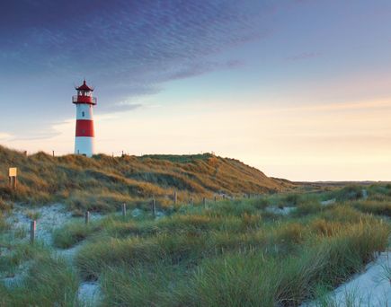 Leuchtturm auf einer Dünenlandschaft mit Gras unter einem bewölkten Himmel bei Sonnenuntergang
