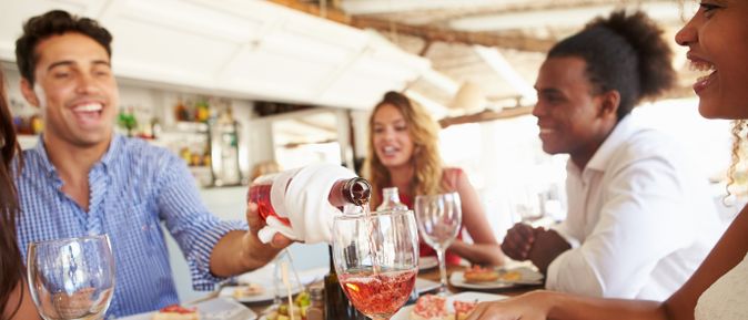 Gruppe von Freunden sitzt fröhlich an einem Tisch im Restaurant, während ein Mann Roséwein in Gläser einschenkt
