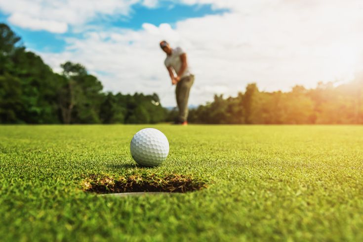 Nahaufnahme eines Golfballs vor dem Loch auf einem gepflegten Grün, im Hintergrund unscharf ein Golfer beim Putten unter sonnigem Himmel