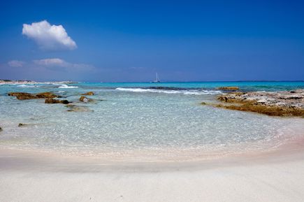 Playa de Levante auf Formentera mit weißem Sandstrand, flachem türkisfarbenem Wasser, Felsen im Meer und einem Segelboot am Horizont