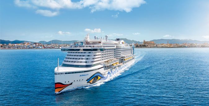 Das Kreuzfahrtschiff AIDAperla fährt auf offenem Meer mit einer Stadt und Bergen im Hintergrund