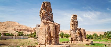 Zwei große, verwitterte Steinstatuen der Memnonkolosse sitzen nebeneinander in einer Wüstenlandschaft mit Bergen und blauem Himmel im Hintergrund