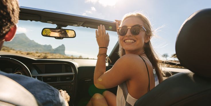 Junges Paar fährt in einem Cabrio bei Sonnenschein, die Frau mit Sonnenbrille lächelt fröhlich in die Kamera