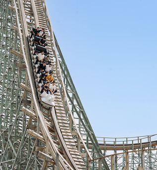 Holzachterbahn mit mehreren Fahrgästen, die eine steile Abfahrt hinunterfahren, vor klarem Himmel.