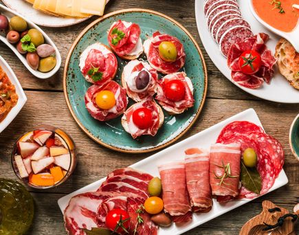 Verschiedene Tapas auf Tellern, darunter Oliven, Tomaten, Schinken, Salami, Brot und ein Gericht mit Tomatensauce, auf einem Holztisch angerichtet.