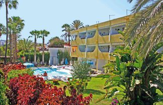 Gelbes Hotelgebäude mit Balkonen neben einem Pool, umgeben von grünen und roten Pflanzen, Palmen und blauem Himmel