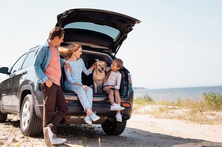 Familie sitzt zusammen mit einem Hund im geöffneten Kofferraum eines geparkten Autos an einer Küstenlandschaft.