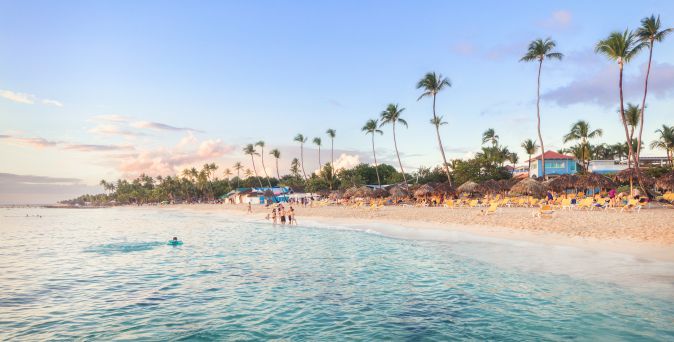 Bayahibe-Strand bei La Romana in der Dominikanischen Republik mit türkisblauem Meer, Palmen, Strohdächern und Liegen entlang des weißen Sandstrands