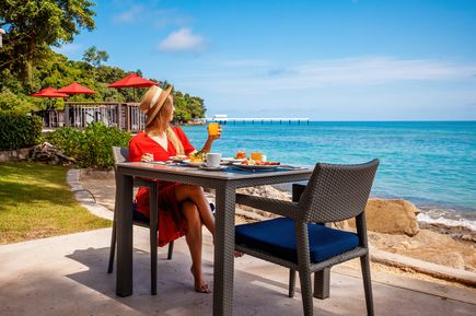 Frau beim Frühstück auf einer Terrasse direkt am Meer mit gedecktem Tisch, Blick auf das türkisfarbene Wasser und eine Küstenlandschaft im Hintergrund