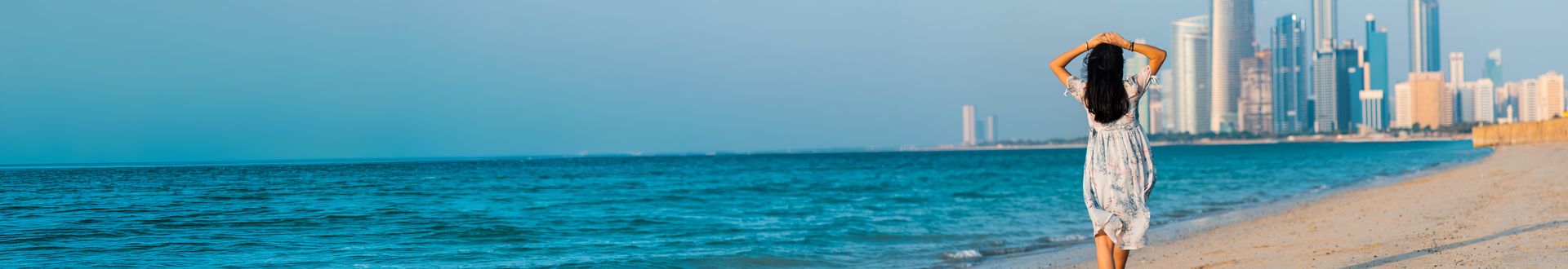 Frau mit langem Kleid steht am Strand und blickt auf die Skyline von Abu Dhabi