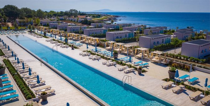 Luftaufnahme eines langgestreckten Pools mit Liegen und Sonnenschirmen, umgeben von modernen Campinghäusern nahe der Küste mit Blick auf das Meer.