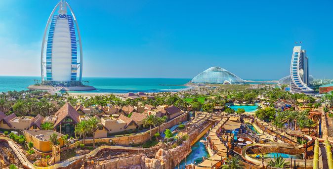 Blick auf den Wild Wadi Wasserpark in Dubai mit Wasserrutschen, Palmen, Bungalows und den markanten Gebäuden Burj Al Arab und Jumeirah Beach Hotel am Meer.