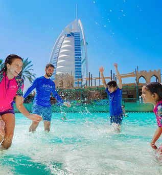 Zwei Kinder und zwei Erwachsene spielen im flachen Wasser des Wild Wadi Wasserparks Dubai, im Hintergrund das markante Burj Al Arab Hotel und Palmen.