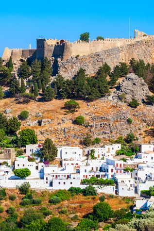 Blick auf die Akropolis von Rhodos auf einem bewaldeten Hügel über einem Dorf mit weißen Häusern und blauem Himmel.