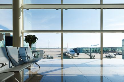 Moderne Wartezone im Flughafenterminal mit Blick auf das Vorfeld, ein angedocktes Passagierflugzeug an der Fluggastbrücke sowie ein startendes Flugzeug im Hintergrund