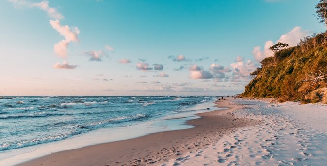 Strand mit Sand und Wellen, rechts bewaldeter Küstenabschnitt unter blauem Himmel mit vereinzelten Wolken