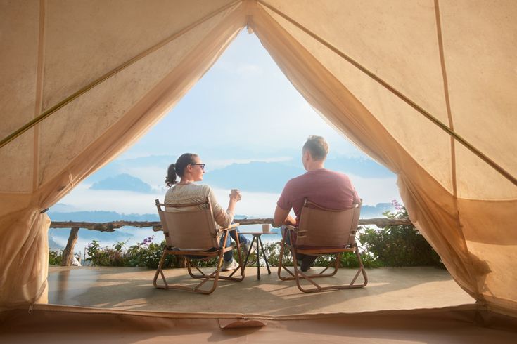 Zwei Personen sitzen auf Stühlen vor einem geöffneten Zelt und blicken über eine Landschaft mit Bergen