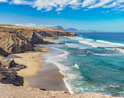 Strand mit goldfarbenem Sand, felsigen Klippen links und blauem Meer mit weißen Wellen rechts, zwei Personen am Strand, bewölkter Himmel