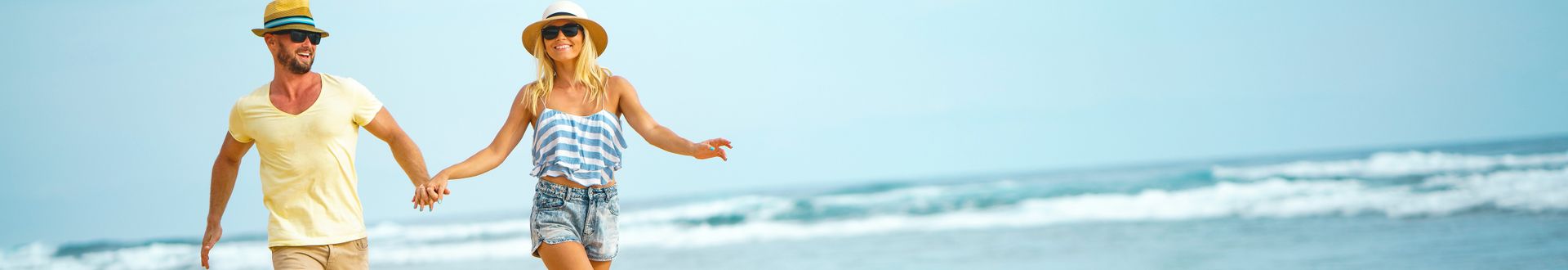 Paar spaziert Hand in Hand am Meer entlang vor einer Kulisse aus blauem Himmel, sanften Wellen und weitläufigem Sandstrand