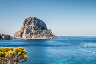 Blick auf die Felseninsel Es Vedrà vor der Küste Ibizas, umgeben vom tiefblauen Meer, mit einem Segelboot im Vordergrund