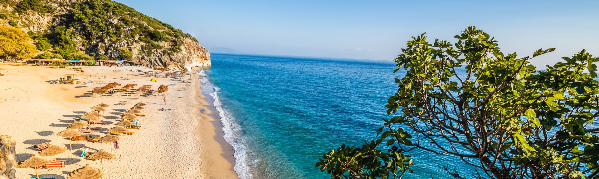 Bild eines Strandabschnittes am blauen Meer, Gebüsch im Vordergrund, ein grüner Hügel im Hintergrund