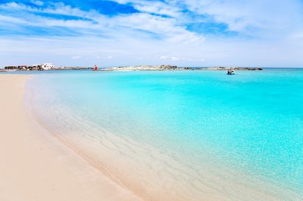 Heller Sandstrand von Es Pujols auf Formentera mit flachem türkisfarbenem Meer und kleinen Gebäuden im Hintergrund