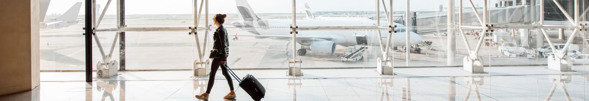 Frau mit Rollkoffer läuft an einer großen Glaswand entlang durch ein modernes Flughafenterminal, während im Hintergrund Flugzeuge auf dem Rollfeld stehen