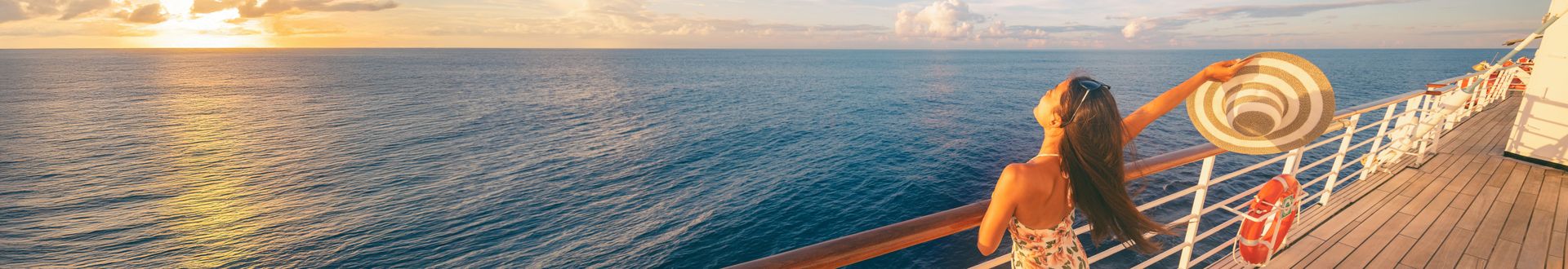 Frau steht auf dem Außendeck eines Kreuzfahrtschiffs und hebt einen Sonnenhut in die Luft, während sich vor ihr der weite Ozean im goldenen Licht des Sonnenuntergangs erstreckt