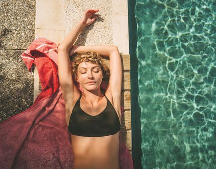 Frau liegt entspannt auf einem Handtuch am Rand eines Pools, lehnt sich mit ausgestreckten Armen zurück und genießt die Sonne neben dem klar schimmernden Wasser