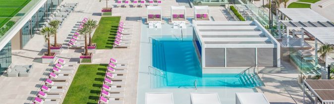 Hotelpool mit Liegen und weißen Sonnendächern, angrenzende Terrasse mit weiteren Liegen, Palmen und Meer im Hintergrund
