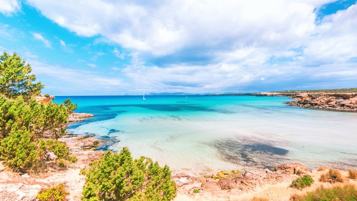 Bucht Cala Saona auf Formentera mit türkisblauem Meer, flachem Sandstrand, felsiger Küste und Booten im Wasser, mediterrane Vegetation unter leicht bewölktem Himmel